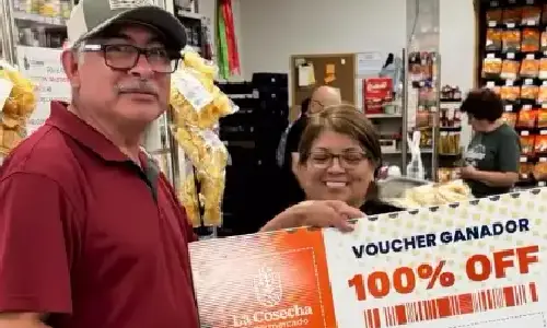 Pareja ganadora en La Cosecha Supermercado Tulsa presume su voucher ganador del 100% de descuento, celebrando junto a su carrito lleno de compras.